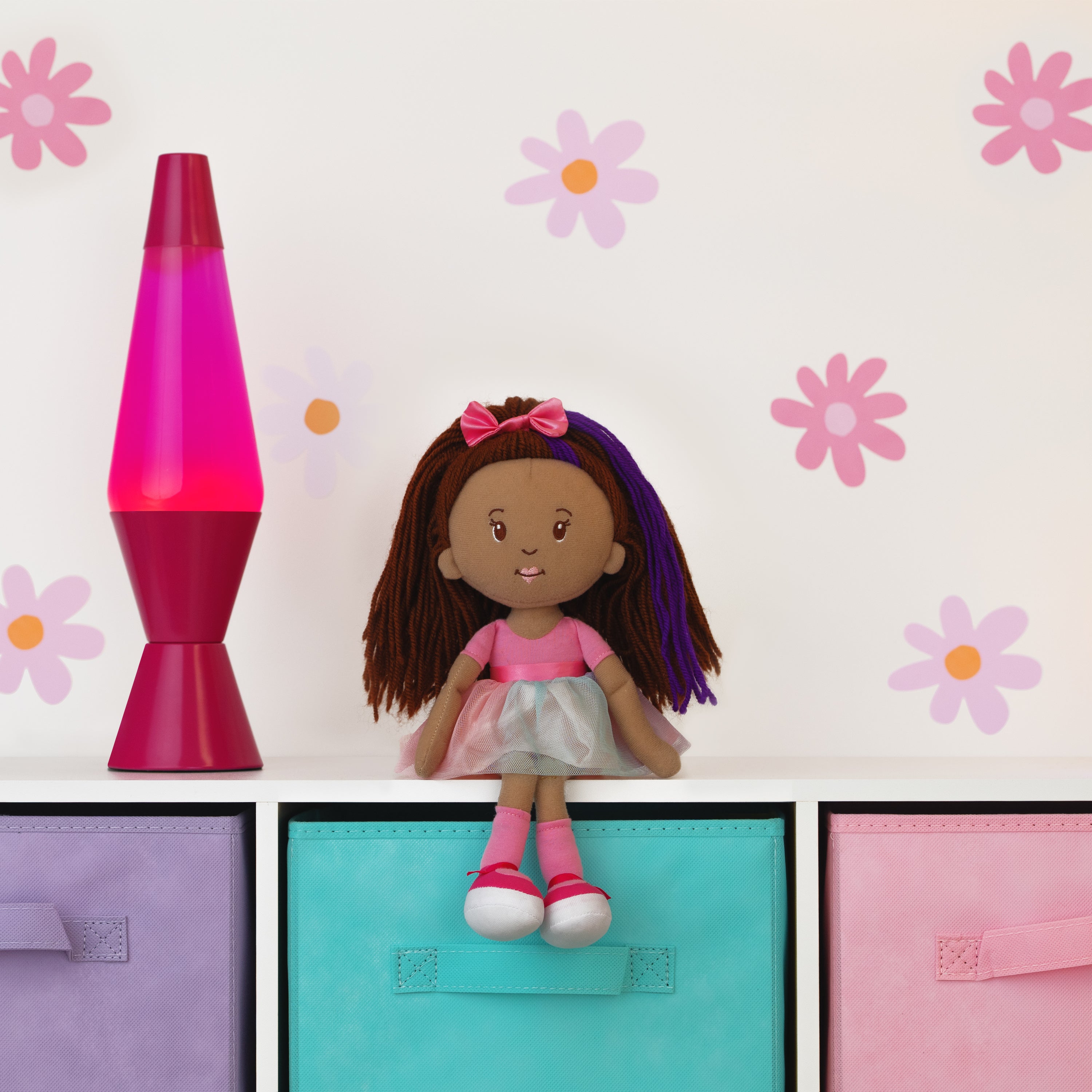 Doll with purple hair and pink bow sitting on a shelf next to a pink lava lamp against a floral wall.