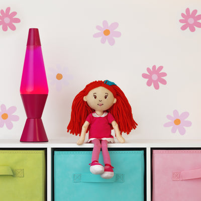 Toy doll with red hair on a shelf next to a pink lava lamp against a wall with flower decals.