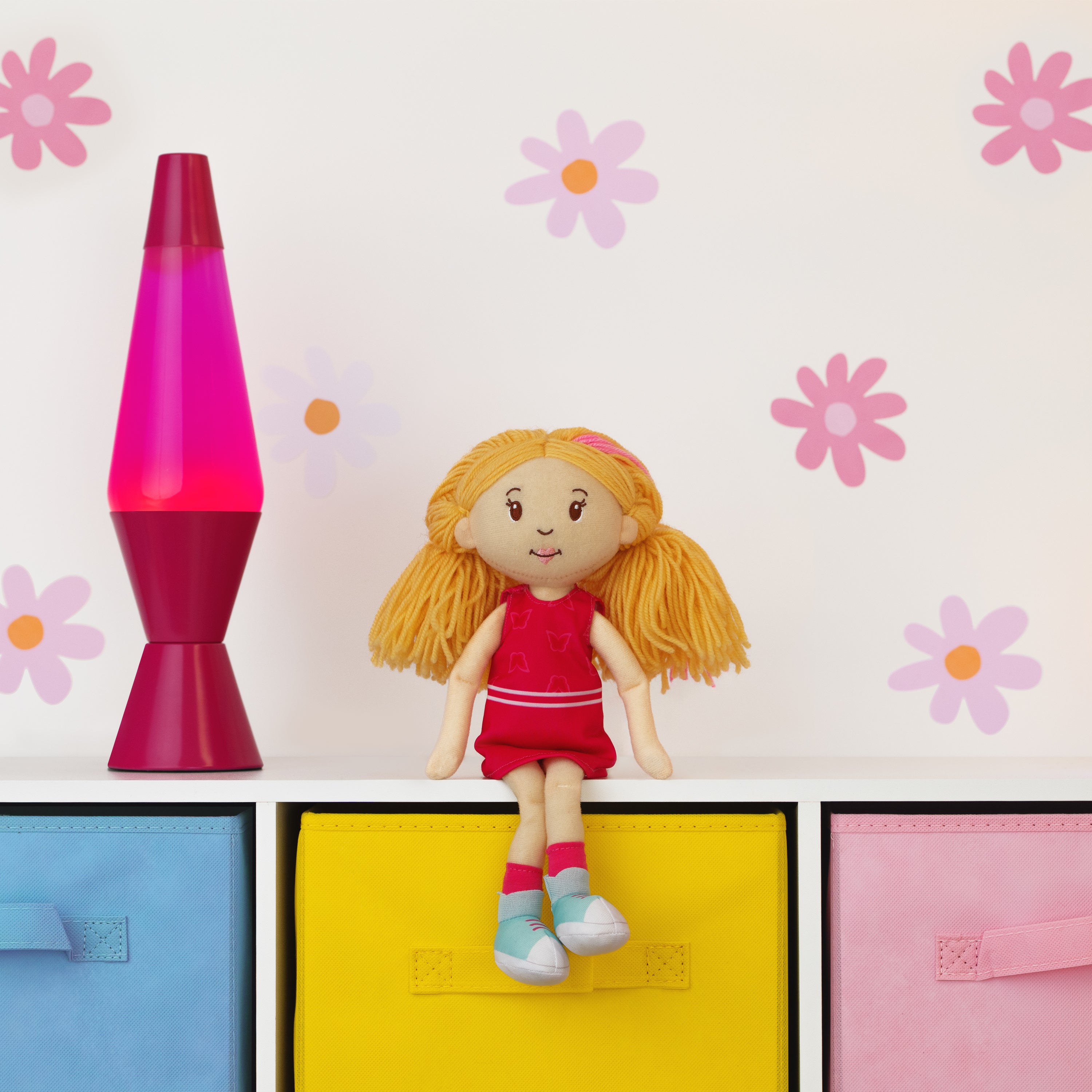 Toy doll with red dress on a shelf with colorful boxes and a pink lava lamp against a floral wall.