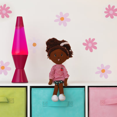 Doll with pink outfit and brown hair sitting on a shelf with a pink lava lamp and colorful boxes against a floral-patterned wall.