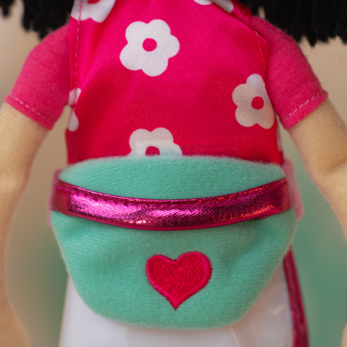Close up of soft doll wearing a pink and white floral shirt with a white athletic skirt and a teal waist bag with a pink heart icon