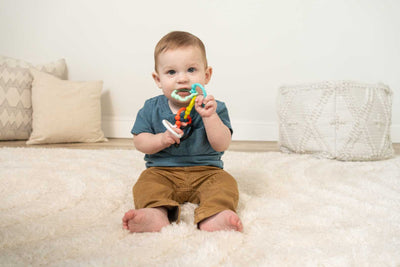 A baby sits on a rug holding multicolored bear-shaped rings in his mouth. 