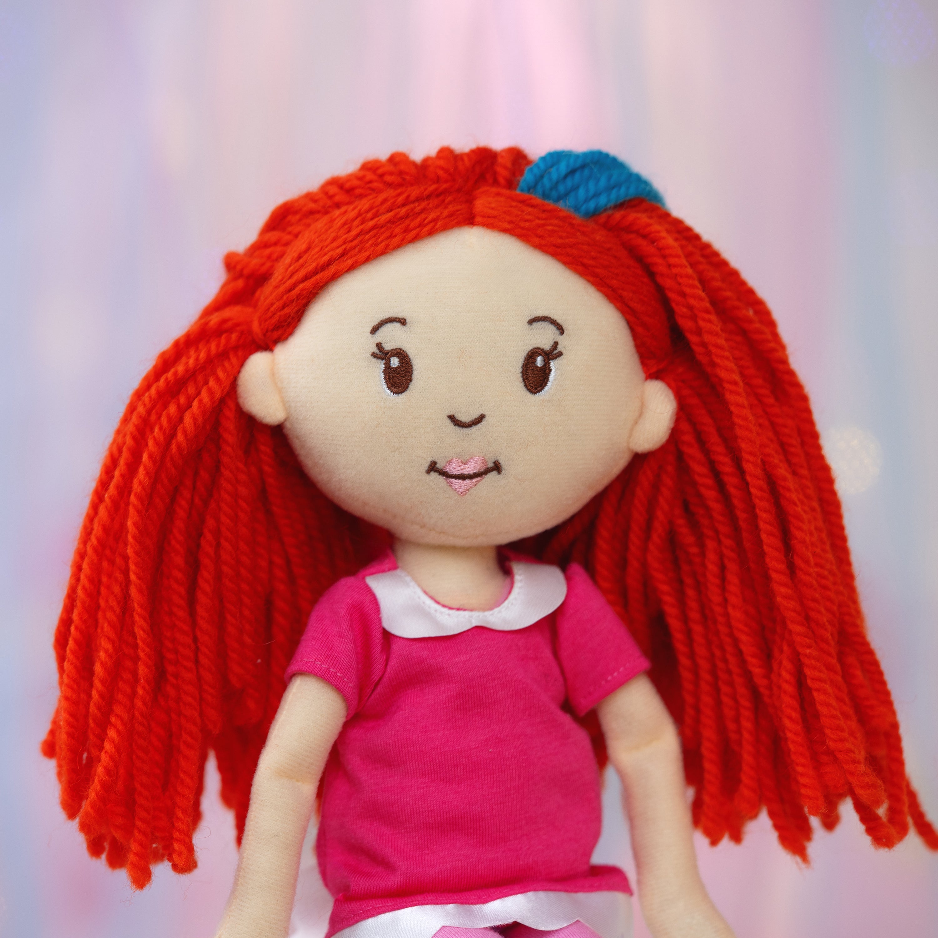 Plush doll with red hair and a pink shirt on a light pink and purple background