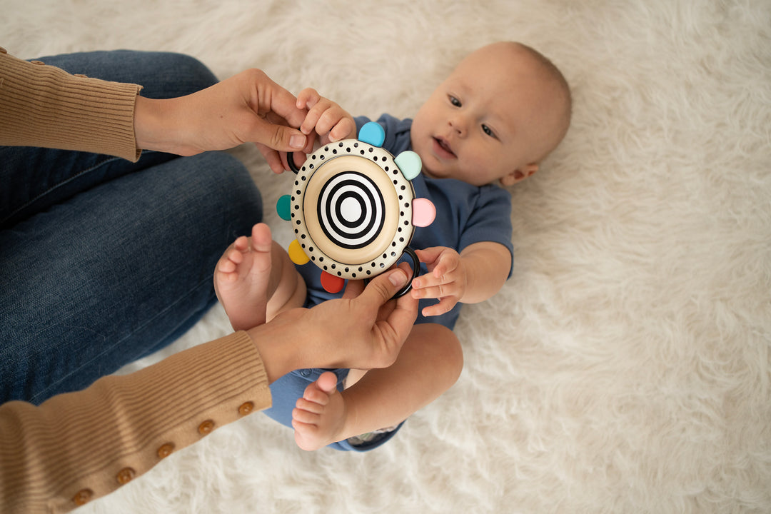 Baby lying on a fluffy surface, holding a colorful rattle toy with help from an adult.