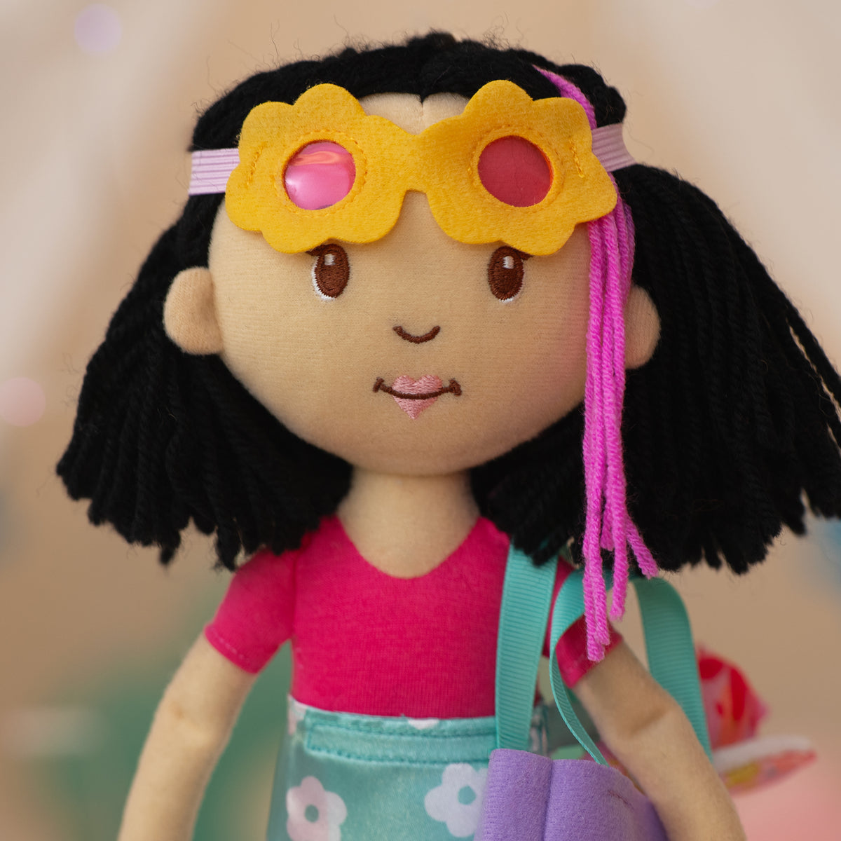 close up of a soft doll wearing yellow flower sunglasses and a pink bodysuit with a blue floral wrap skirt.