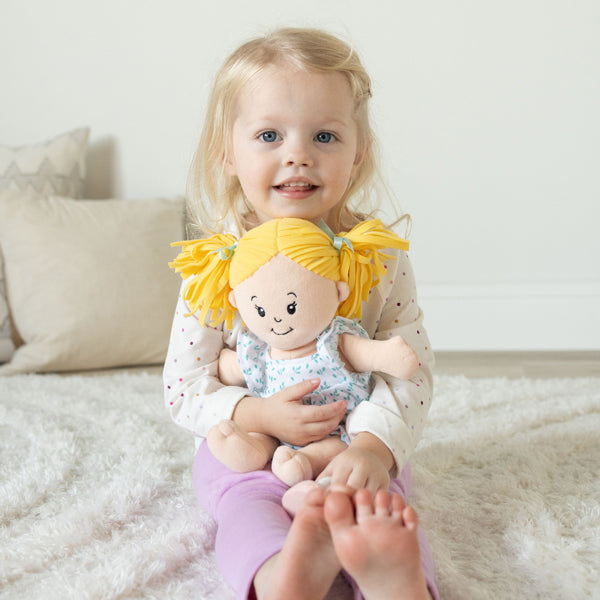 Young girl sitting on a soft rug, smiling and hugging a blonde-haired plush doll.