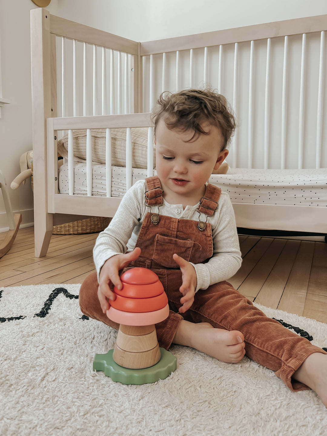Toddler in overalls playing with a colorful stacking toy in a nursery.