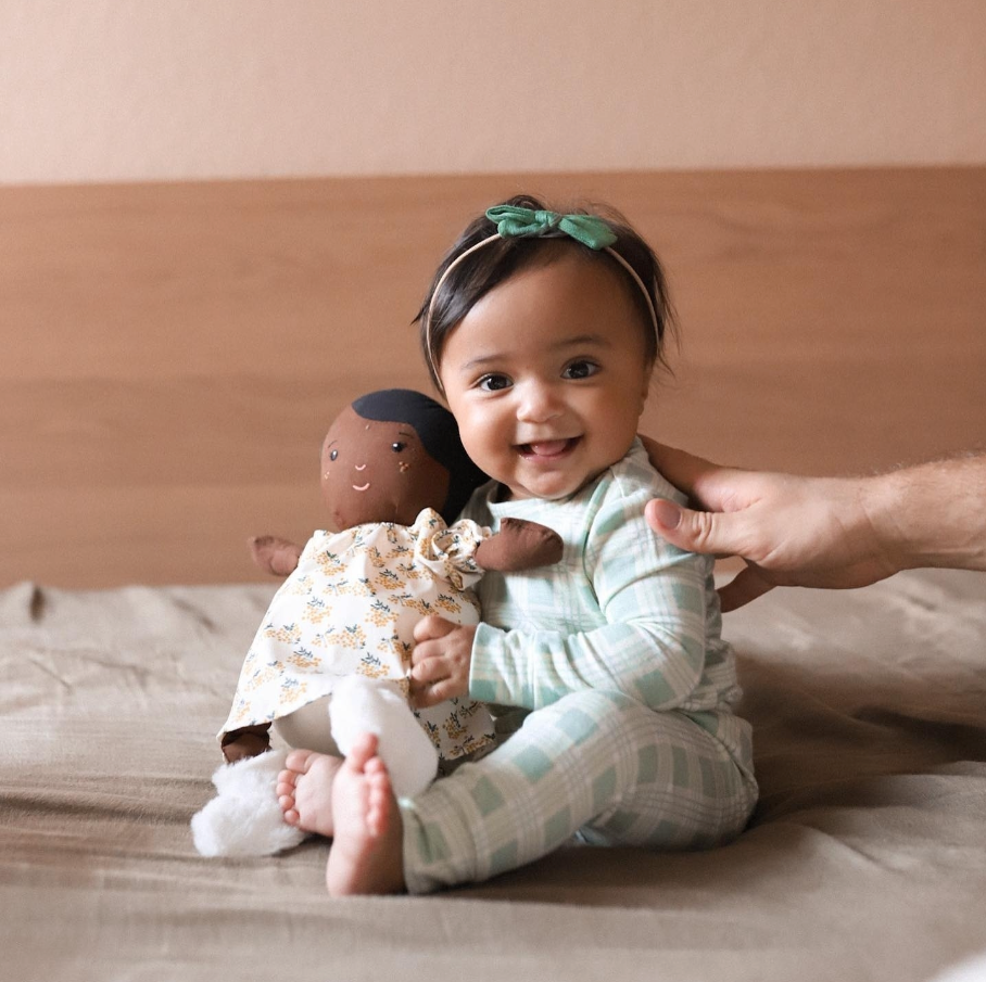 Baby smiling and holding a doll while being gently supported by an adult hand.