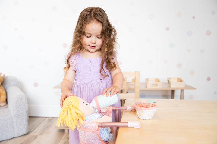 A little girl feeds her doll a bottle while it sits in a pink doll table chair.