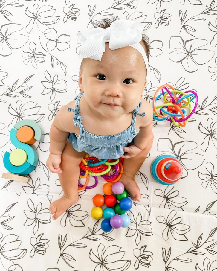 Baby in a blue romper with a large white bow, surrounded by colorful toys on a floral-patterned mat.