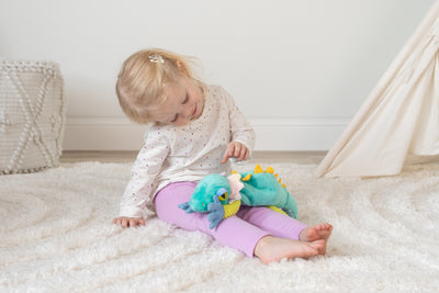 A toddler sits on a rug smiling down at a green plush lizard on her lap.