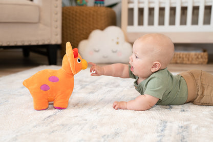 A baby lays on his tummy on a rug reaching for a plush orange triceratops with purple spots. 