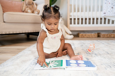 A toddler sits on a rug in a bedroom with My First Photobook open to a green and blue page on the floor in front of her. 