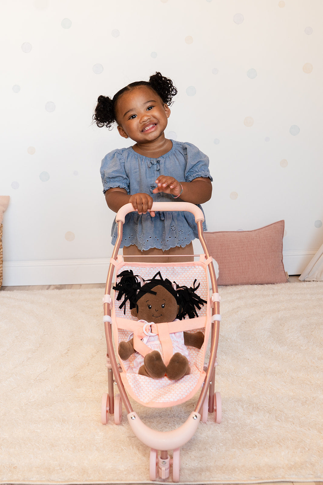A little girl smiles wide while holding onto a pink doll stroller with a doll sitting inside.