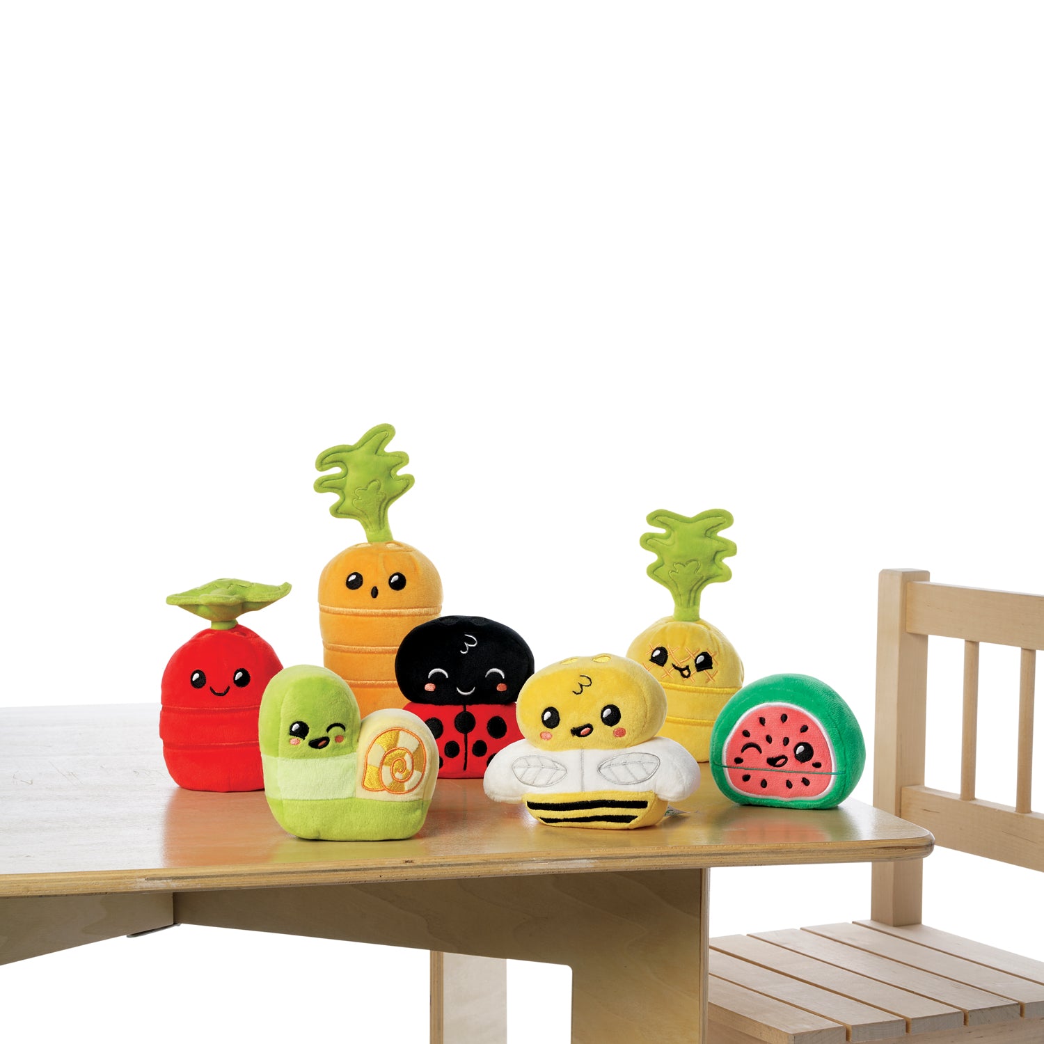A group of colorful plush toys shaped like fruits, vegetables, and insects, including a carrot, bee, snail, watermelon, tomato, and ladybug, displayed on a wooden table and chair set.