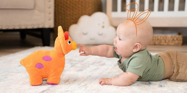 Baby boy with hand-draw bunny ears on head, laying on tummy facing and reaching towards orange triceratops dino in a nursery room scene.