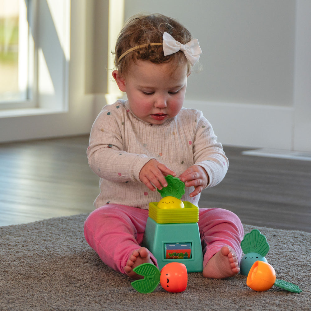 Toddler sitting on a carpeted floor, playing with a colorful vegetable-themed stacking toy, focusing on placing a green leafy piece into a yellow container.