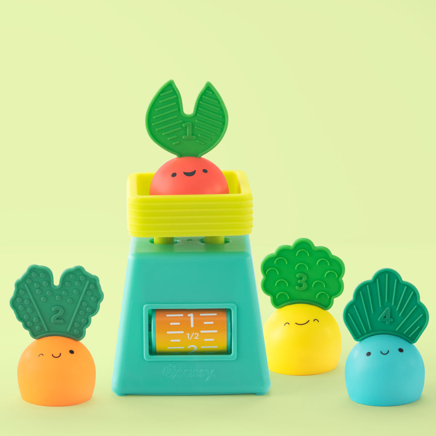 Colorful vegetable-themed stacking toy set with smiling carrots, broccoli, and leafy greens displayed around a teal and yellow toy scale on a light green background.