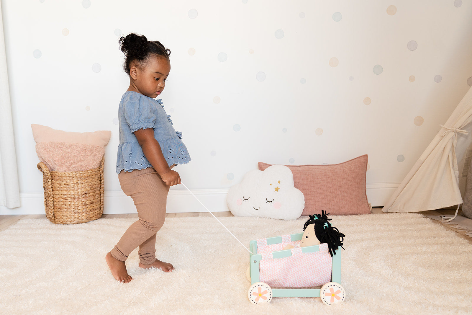 A toddler holds the pull cord of a blue and pink wooden doll wagon with a doll sitting inside.