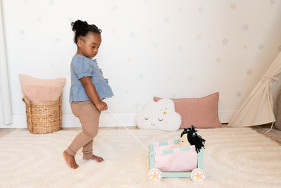 A toddler holds the pull cord of a blue and pink wooden doll wagon with a doll sitting inside.