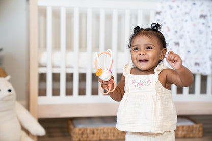 A smiling toddler holds a bunny shaped rattle with a white center and pink looping plastic cords with orange and pink beads